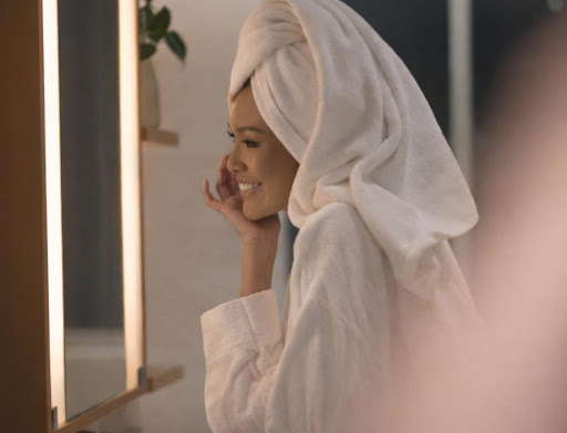 A serene, high-fashion editorial photograph of a woman with her hair in a white towel, smiling at her reflection in a well-lit vanity mirror at Brittany Hotel BGC. The atmosphere is calm and luxurious, emphasizing morning wellness and rejuvenation.