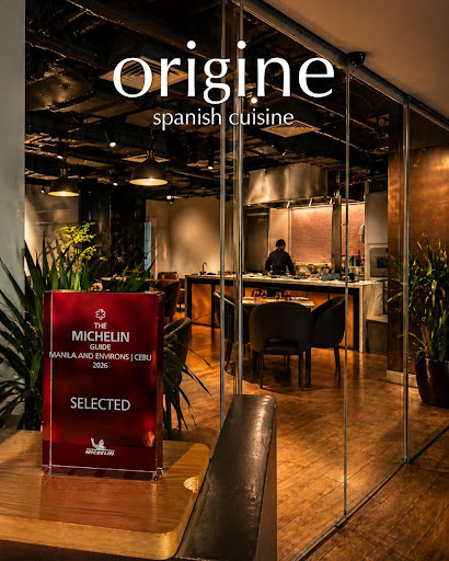 An intimate, high-end Spanish restaurant interior (Origine BGC). Warm ambient lighting, dark wood tables, elegant table settings with wine glasses. A quiet corner table prepared for for a romantic dinner. Sophisticated and inviting atmosphere. Professional food and interior photography.
