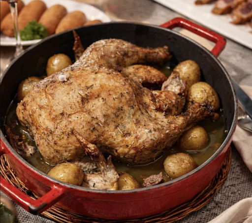 A roasted whole chicken with golden-brown skin, surrounded by small roasted potatoes in a red cast-iron pan with a light broth or sauce.