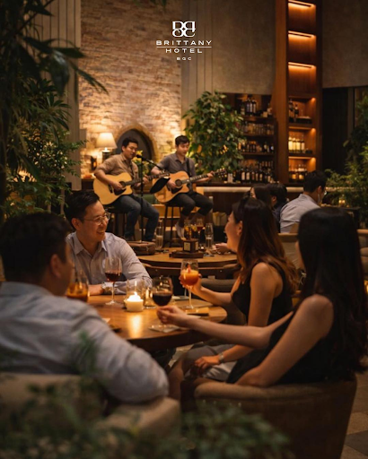 A warm, atmospheric dining scene at Brittany Hotel BGC featuring live acoustic musicians performing in the background while guests enjoy wine and conversation in a sophisticated, dimly lit setting with rich textures and golden ambient lighting.