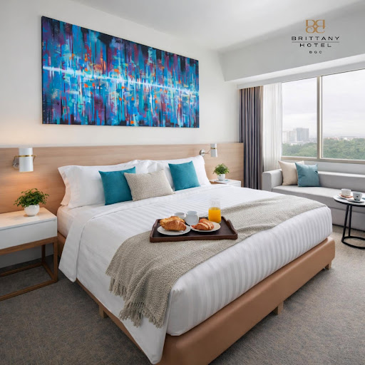A minimalist and airy deluxe hotel room interior with soft natural lighting streaming through large windows. The room features a plush king-sized bed with white linens, sand-colored walls, and subtle sage green accents. The furniture is clean-lined and modern, crafted from light-toned wood. The image captures a sense of profound tranquility and stillness, typical of a high-end wellness retreat.