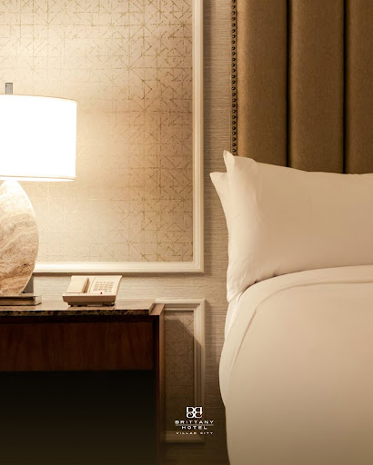 A minimalist and high-end hotel room interior featuring a large king-size bed with crisp white linens and a plush charcoal throw. The room is bathed in soft, natural morning light filtering through sheer curtains, creating a serene and exclusive atmosphere. The design follows a quiet luxury aesthetic with neutral tones, clean lines, and premium textures. No people are present, emphasizing the solitude and calm of a solo travel experience.