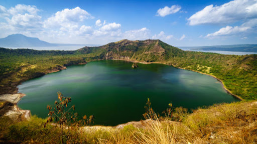 A cinematic, wide-angle landscape of Taal Volcano in the Philippines, viewed from a high vantage point in Tagaytay. The scene is bathed in the soft, warm golden hour light of a tropical afternoon, highlighting the deep forest greens of the surrounding foliage and the serene blue of the lake. The overall aesthetic is one of quiet luxury and natural grandeur, aligned with a premium travel magazine editorial style.