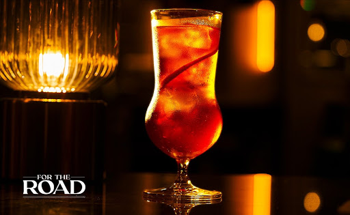 A close-up of a perfectly crafted cocktail with a garnish of orange peel sitting on a dark marble bar counter. The background shows a sophisticated, softly lit bar with rows of premium bottles and a warm, amber glow from pendant lights. The aesthetic is moody and elegant, capturing a quiet late-night moment of luxury and shared connection.