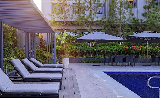 A serene infinity pool reflecting a vibrant sunset of orange, pink, and deep violet. The surrounding deck is made of soft-clay colored stone with elegant lounge chairs. The lighting is low and atmospheric, emphasizing the tranquil water surface and the silhouettes of tall trees in the distance, evoking a peaceful, romantic evening in a high-end luxury resort.