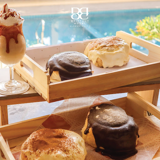 A beautifully set hotel cafe table with coffee and Filipino delicacies under warm, golden afternoon sunlight. The scene is elegant and inviting, featuring premium ceramic tableware and soft-focus backgrounds of a lush tropical garden visible through large glass windows.